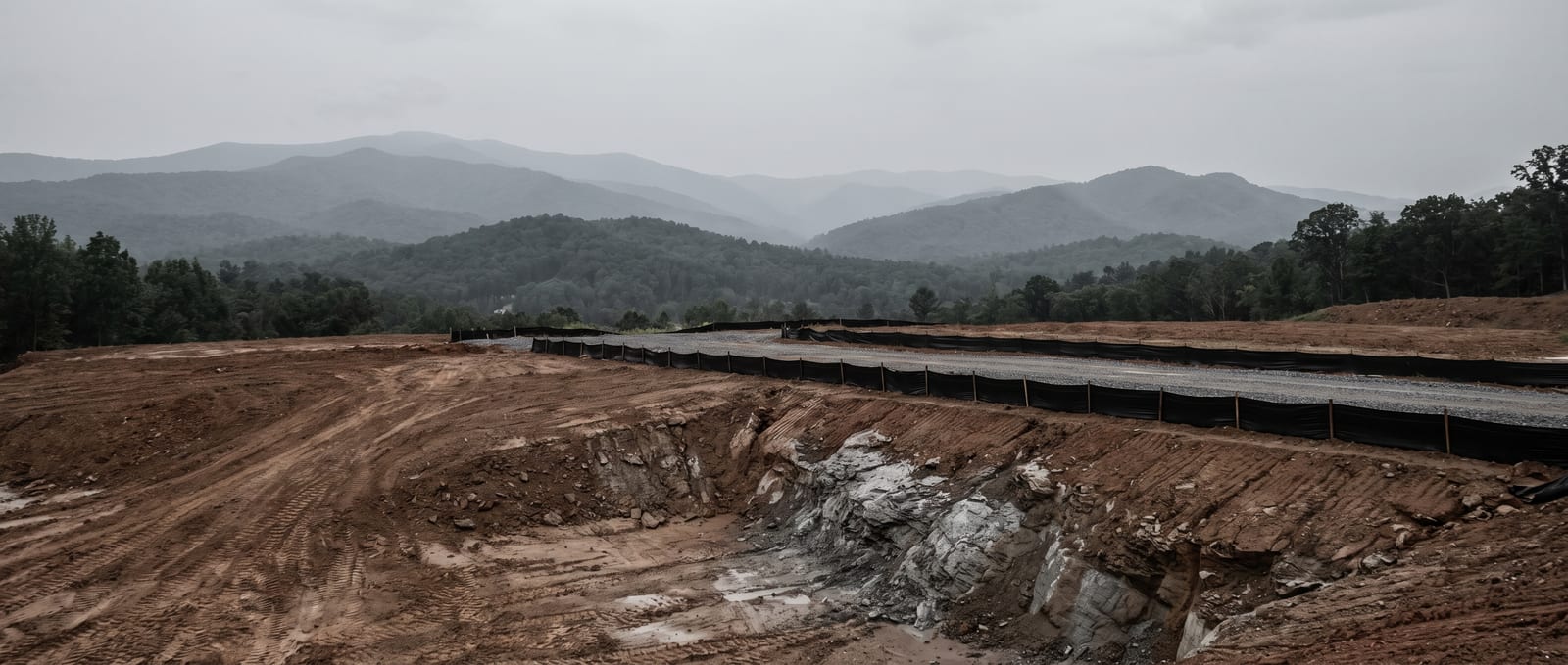 A sloped, partly cleared Buncombe County building lot in the Western North Carolina mountains, disturbed soil in the foreground with wooded ridgelines behind