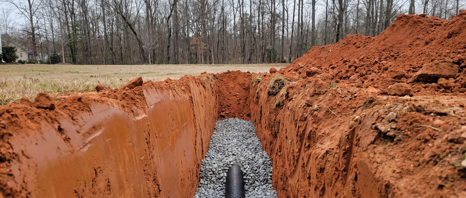 Open French drain trench cut into NC Piedmont red clay, glazed trench wall visible on one side and washed stone with perforated pipe being placed in the base