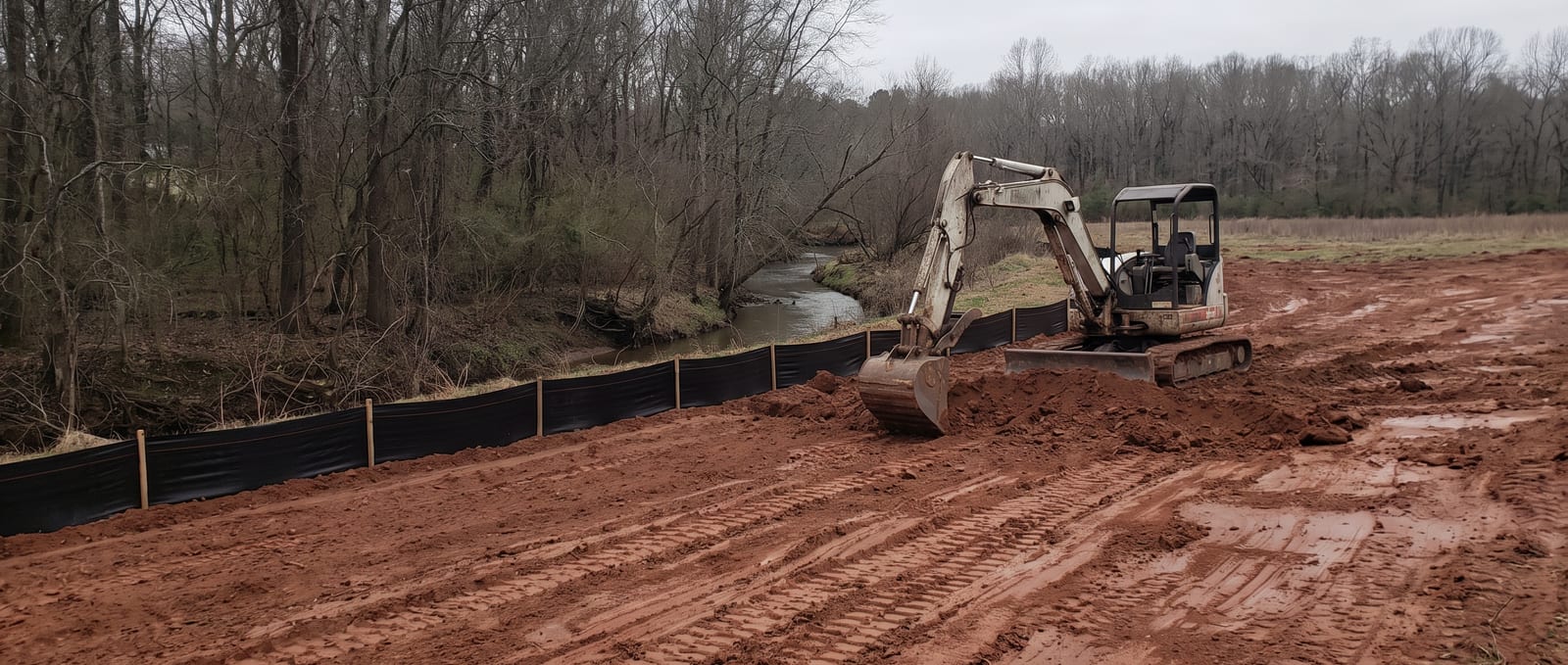 A compact excavator working red-clay ground on a Greensboro-area lot, silt fence staked along a wooded stream buffer at the boundary, under flat overcast Piedmont light