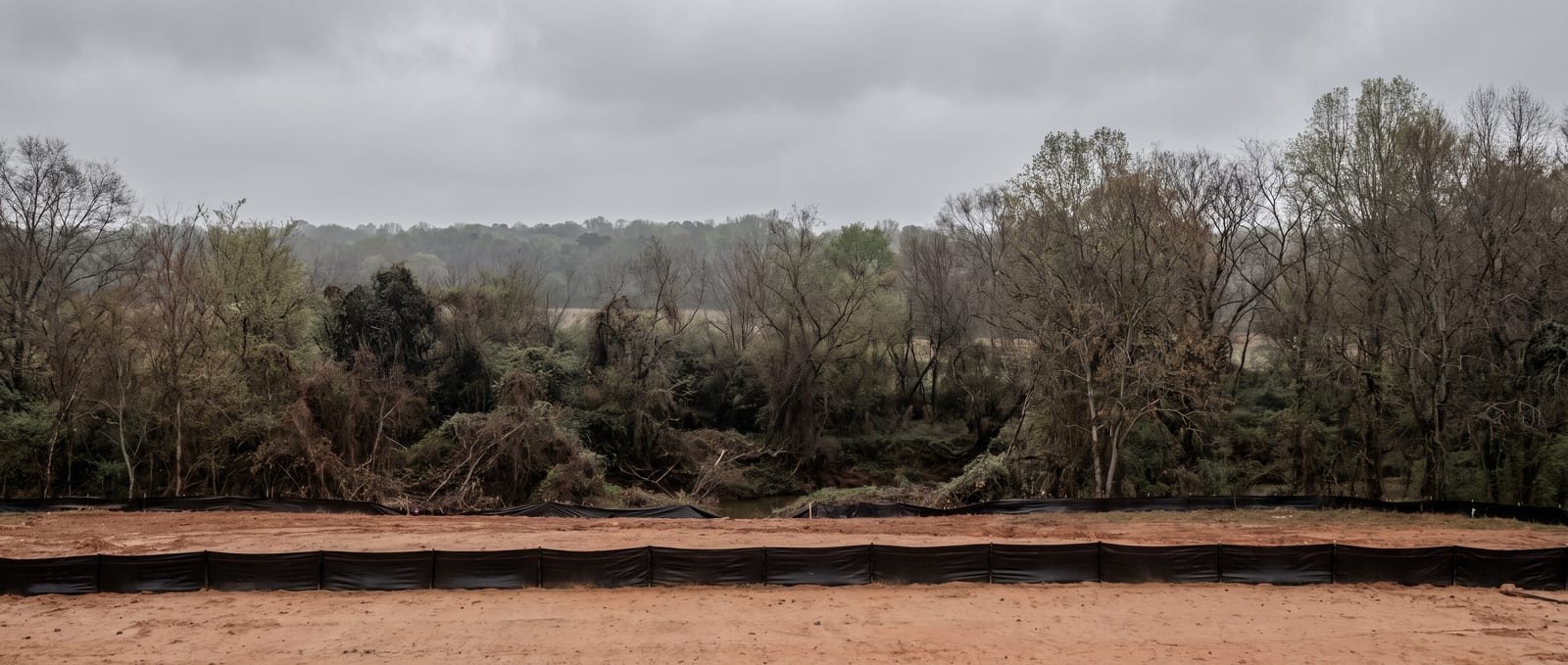 A graded red-clay residential lot in Mecklenburg County beside a wooded stream buffer, silt fence marking the no-disturbance line under an overcast Piedmont sky